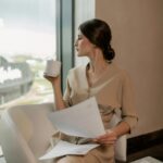 pexels-photo-8134238-8134238 Sophisticated woman in beige dress holding a cup of coffee and papers, seated indoors beside a window.